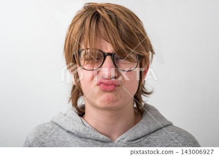Young person wearing glasses and a grey hoodie is expressing dislike or dissatisfaction, squinting eyes and scrunching lips in a pouting grimace against a white background Young person wearing glasses and a grey hoodie is expressing dislike or dissatisfaction, squinting eyes and scrunching lips in a pouting grimace against a white background 143006927