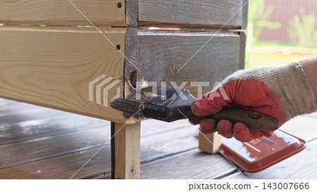 Hand with brush painting wooden planks with brown protective oil finish close up. Wood staining process. Person applying dark varnish coat on timber board outdoors. House remodeling and DIY renovation Hand with brush painting wooden planks with brown protective oil finish close up. Wood staining process. Person applying dark varnish coat on timber board outdoors. House remodeling and DIY renovation 143007666