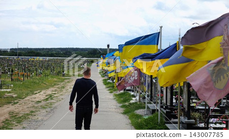 Mourning man go among countless graves of ukrainian soldiers with national flags at military cemetery in Kharkiv. Scene reflects personal sorrow and nation collective loss caused by russian aggression Mourning man go among countless graves of ukrainian soldiers with national flags at military cemetery in Kharkiv. Scene reflects personal sorrow and nation collective loss caused by russian aggression 143007785