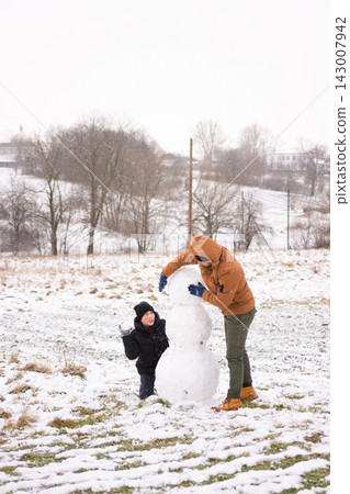 A father and son build a snowman together in a snowy field. The boy watches as his dad shapes the snow. A father and son build a snowman together in a snowy field. The boy watches as his dad shapes the snow. 143007942