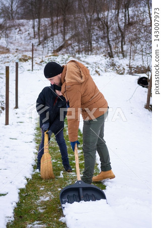 Father and son teamwork: clearing snow from a pathway using a shovel and broom. Winter cleanup. Father and son teamwork: clearing snow from a pathway using a shovel and broom. Winter cleanup. 143007973