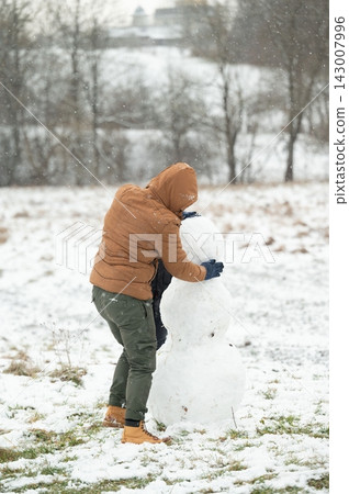 Building a snowman on a snowy day. A person in a brown jacket carefully packs snow onto the snowman's body. Building a snowman on a snowy day. A person in a brown jacket carefully packs snow onto the snowman's body. 143007996