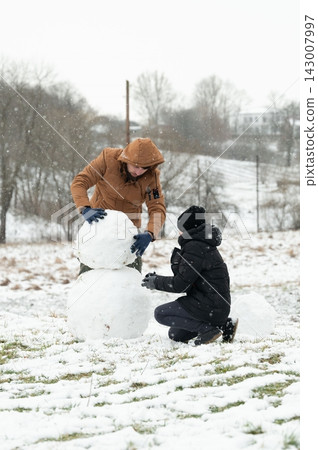 Two individuals collaborate on building a snowman in a snowy landscape, enjoying a winter day. Two individuals collaborate on building a snowman in a snowy landscape, enjoying a winter day. 143007997