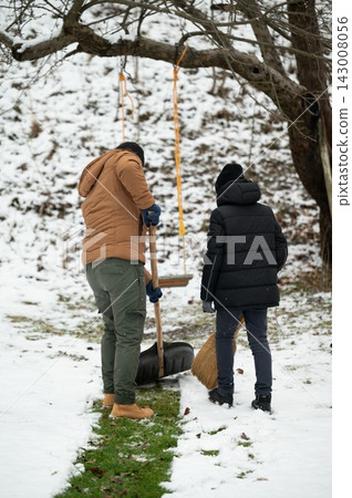 Two individuals collaborate on clearing snow from a pathway using a shovel and a broom, a winter chore. Two individuals collaborate on clearing snow from a pathway using a shovel and a broom, a winter chore. 143008056
