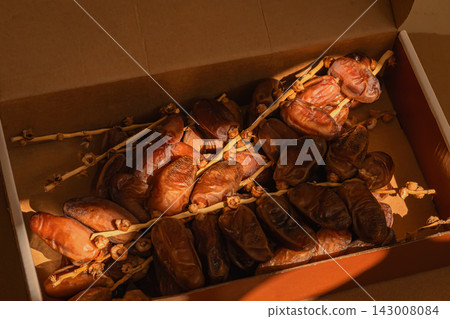 Dried Dates on Palm Branches in Cardboard Box. Ripe dried dates attached to palm stems inside cardboard box in warm natural sunlight. Natural food still life, rich brown tones, rustic harvest texture Dried Dates on Palm Branches in Cardboard Box. Ripe dried dates attached to palm stems inside cardboard box in warm natural sunlight. Natural food still life, rich brown tones, rustic harvest texture 143008084