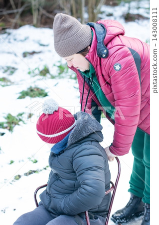 A person helps a child sit on a small sled in a snowy woodland setting. A person helps a child sit on a small sled in a snowy woodland setting. 143008111