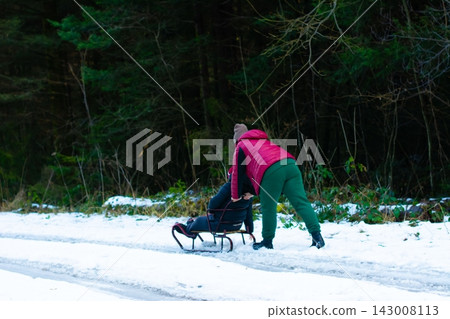 A person pulls a sled carrying another person through a snowy forest path. Winter fun in the woods. A person pulls a sled carrying another person through a snowy forest path. Winter fun in the woods. 143008113