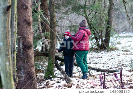 A parent and child explore a snowy winter forest, examining a tree. 143008125