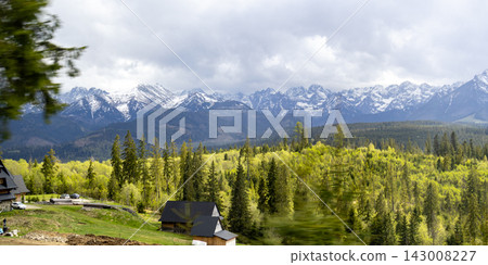 Panoramic view of snow covered High Tatras mountains above green forest and rolling countryside near Zakopane. Scenic alpine landscape under dramatic spring clouds in Poland. 143008227