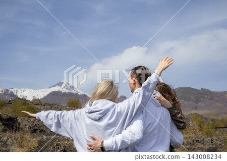 Back view of family standing with open arms and enjoying of the dramatic volcanic landscape of Mount Etna. Concept of freedom, adventure and connection with nature. Back view of family standing with open arms and enjoying of the dramatic volcanic landscape of Mount Etna. Concept of freedom, adventure and connection with nature. 143008234