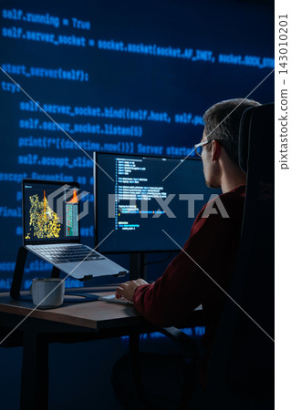 Coder works at desk in a dark office with programming code lines running behind Coder works at desk in a dark office with programming code lines running behind 143010201