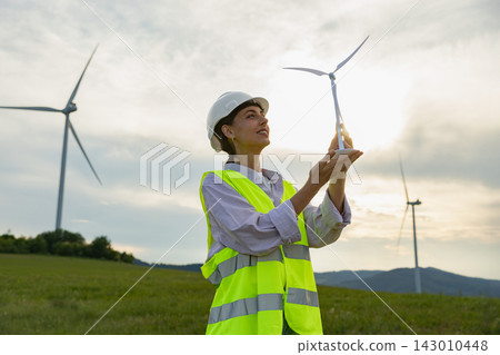 A Caucasian female professional in a safety vest and hard hat examines a miniature wind turbine model with a large wind farm in the background A Caucasian female professional in a safety vest and hard hat examines a miniature wind turbine model with a large wind farm in the background 143010448