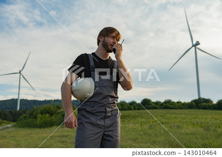 A man in overalls and a black t-shirt holds a white hard hat while speaking into a walkie-talkie with wind turbines in the background 143010464