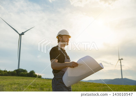 A man in work overalls and a hard hat stands in a grassy field holding rolled-up plans with wind turbines in the background during golden hour A man in work overalls and a hard hat stands in a grassy field holding rolled-up plans with wind turbines in the background during golden hour 143010547