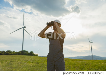 A man in work clothes surveys a field with wind turbines under a cloudy sky, symbolizing renewable energy and observation A man in work clothes surveys a field with wind turbines under a cloudy sky, symbolizing renewable energy and observation 143010579