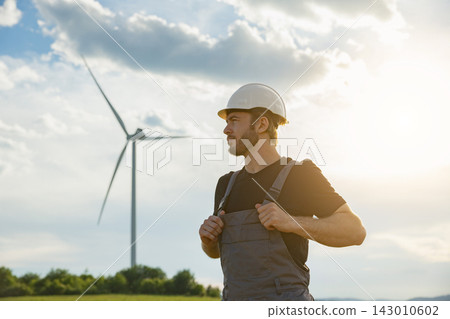A man in work clothes stands in a field with a wind turbine behind him under a bright sky with clouds A man in work clothes stands in a field with a wind turbine behind him under a bright sky with clouds 143010602