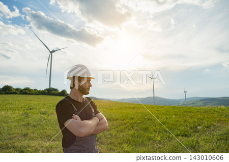 A Caucasian engineer wearing a hard hat and overalls stands with arms crossed in a grassy field overlooking wind turbines A Caucasian engineer wearing a hard hat and overalls stands with arms crossed in a grassy field overlooking wind turbines 143010606