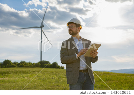 A Caucasian man wearing a hard hat and glasses holds a tablet while standing in a grassy field with a wind turbine in the background A Caucasian man wearing a hard hat and glasses holds a tablet while standing in a grassy field with a wind turbine in the background 143010631