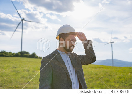 A Caucasian engineer wearing a hard hat and suit stands in a grassy field with wind turbines in the background on a sunny day A Caucasian engineer wearing a hard hat and suit stands in a grassy field with wind turbines in the background on a sunny day 143010686