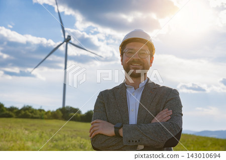 A Caucasian engineer wearing a hard hat and glasses stands in a field with a wind turbine behind him A Caucasian engineer wearing a hard hat and glasses stands in a field with a wind turbine behind him 143010694
