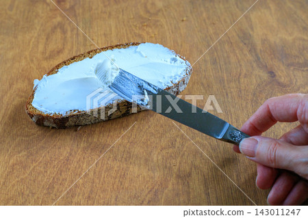 Hand Spreading White Soft Cheese on Rye Bread Slice with Knife on Wooden Table. Morning Breakfast Concept Hand Spreading White Soft Cheese on Rye Bread Slice with Knife on Wooden Table. Morning Breakfast Concept 143011247