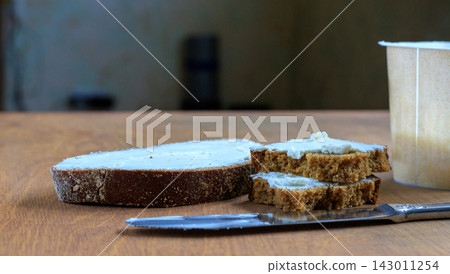 Rye Bread Sandwiches with Soft White Spread, Knife and Dairy Container on Wooden Surface Rye Bread Sandwiches with Soft White Spread, Knife and Dairy Container on Wooden Surface 143011254