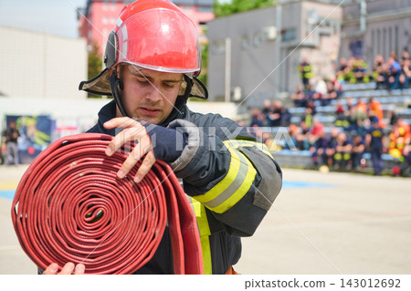 Firefighter Rolling a Red Fire Hose During Training Exercise Wearing Helmet and Protective Gear 143012692