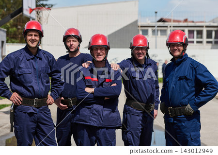 Smiling Firefighters Team in Red Helmets and Navy Uniforms Standing Together Outside Fire Station Smiling Firefighters Team in Red Helmets and Navy Uniforms Standing Together Outside Fire Station 143012949