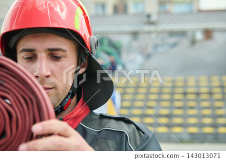 Young Firefighter In Red Helmet Rolling Hose During Outdoor Training Drill At Stadium Young Firefighter In Red Helmet Rolling Hose During Outdoor Training Drill At Stadium 143013071