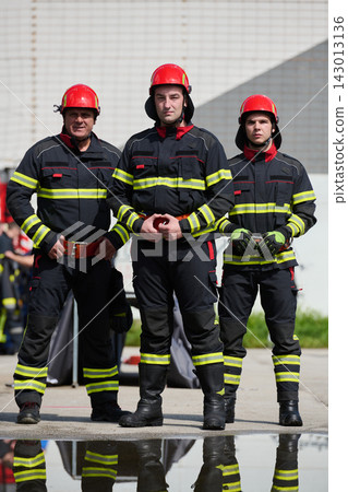 Three Firefighters Standing in Uniform With Red Helmets and Reflective Stripes Ready for Rescue Duty 143013136