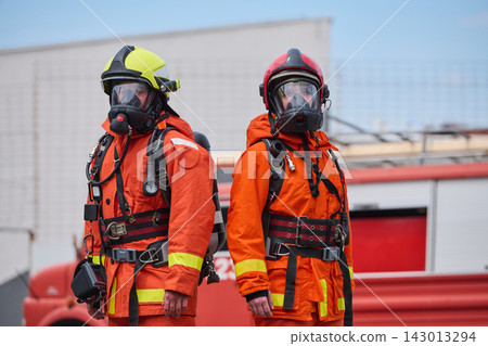 Two Firefighters In Orange Protective Gear With Respirator Masks And Helmets Standing By Fire Truck Two Firefighters In Orange Protective Gear With Respirator Masks And Helmets Standing By Fire Truck 143013294