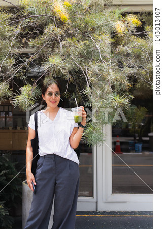 Stylish Asian woman wearing sunglasses holding an iced matcha green tea while posing under a flowering tree outside a cafe. Stylish Asian woman wearing sunglasses holding an iced matcha green tea while posing under a flowering tree outside a cafe. 143013407