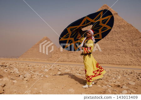 Proud folk performer in embroidered yellow galabiya and white headwrap holding bold geometric parasol at the Giza pyramids in Egypt ideal for travel Proud folk performer in embroidered yellow galabiya and white headwrap holding bold geometric parasol at the Giza pyramids in Egypt ideal for travel 143013409