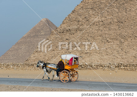 Authentic horse drawn carriage with orange wooden wheels and white canopy moving in profile past the towering limestone base wall of the Giza pyramids 143013662