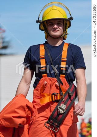 Confident Young Firefighter in Yellow Helmet and Orange Protective Gear Holding Jacket Outdoors 143013680