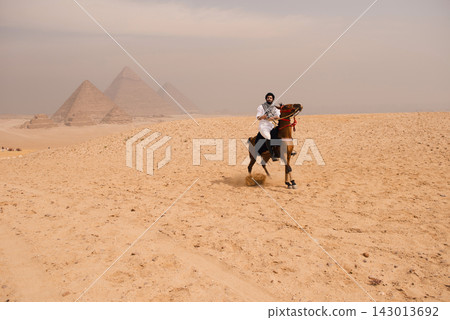 Fearless Arab horseman in white shirt and keffiyeh rides horse at full gallop across the sandy Giza desert plateau with the Great Pyramids rising in Fearless Arab horseman in white shirt and keffiyeh rides horse at full gallop across the sandy Giza desert plateau with the Great Pyramids rising in 143013692