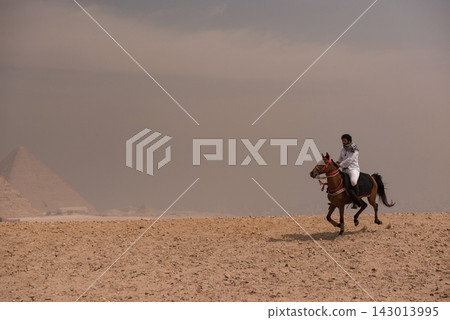 Bold Arab horseman in traditional white thobe and keffiyeh galloping at full speed across a sandy desert plateau near the ancient Giza pyramids under Bold Arab horseman in traditional white thobe and keffiyeh galloping at full speed across a sandy desert plateau near the ancient Giza pyramids under 143013995