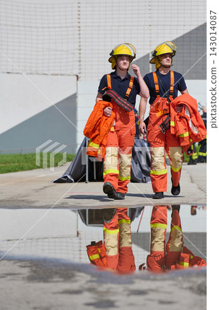 Two Firefighters Walking in Orange Turnout Gear With Yellow Helmets and Reflection in Puddle Two Firefighters Walking in Orange Turnout Gear With Yellow Helmets and Reflection in Puddle 143014087