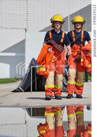 Two Firefighters Walking With Gear And Helmets During Training Exercise With Reflective Puddle Two Firefighters Walking With Gear And Helmets During Training Exercise With Reflective Puddle 143014113