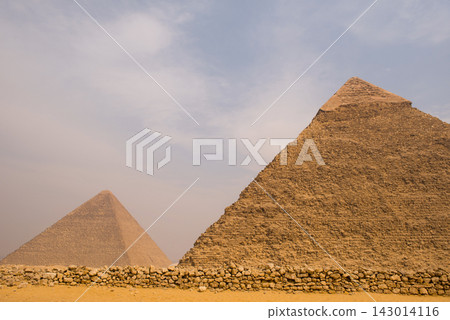 Staggered two pyramid view at Giza plateau with weathered rubble wall in sandy foreground under diffused overcast sky ideal for Egypt travel and Staggered two pyramid view at Giza plateau with weathered rubble wall in sandy foreground under diffused overcast sky ideal for Egypt travel and 143014116