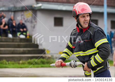 Firefighter In Protective Uniform And Red Helmet Holding Hose During Training Drill With Team In Background Firefighter In Protective Uniform And Red Helmet Holding Hose During Training Drill With Team In Background 143014130