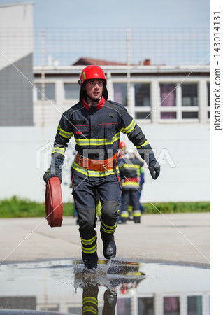 Firefighter Running With Fire Hose During Emergency Drill Wearing Protective Gear And Red Helmet Firefighter Running With Fire Hose During Emergency Drill Wearing Protective Gear And Red Helmet 143014131