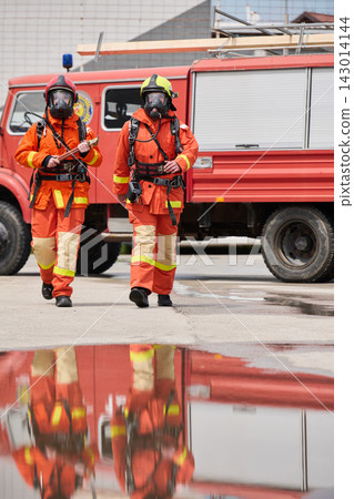 Two Firefighters in Full Protective Gear Walking in Front of Red Fire Truck With Reflection Two Firefighters in Full Protective Gear Walking in Front of Red Fire Truck With Reflection 143014144