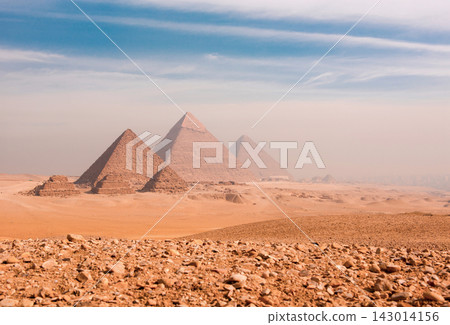 Majestic giza pyramids aligned across vast rocky Egyptian desert plateau under atmospheric hazy blue sky ideal for travel editorial and world history Majestic giza pyramids aligned across vast rocky Egyptian desert plateau under atmospheric hazy blue sky ideal for travel editorial and world history 143014156