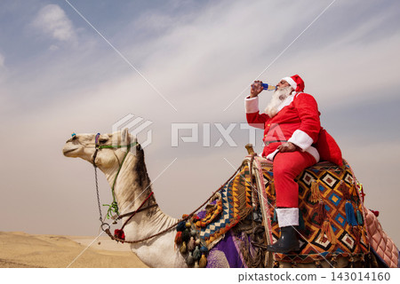 Santa Claus riding a colorfully decorated camel and drinking from a bottle in a vast sandy desert under overcast sky humorous Christmas holiday Santa Claus riding a colorfully decorated camel and drinking from a bottle in a vast sandy desert under overcast sky humorous Christmas holiday 143014160