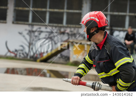 Firefighter in Red Helmet Holding Hose During Urban Fire Drill Near Abandoned Building Firefighter in Red Helmet Holding Hose During Urban Fire Drill Near Abandoned Building 143014162