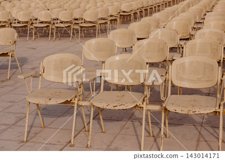 Rows of empty weathered cream metal chairs with rust detail arranged on a tiled plaza at an open air outdoor venue before a public event or concert Rows of empty weathered cream metal chairs with rust detail arranged on a tiled plaza at an open air outdoor venue before a public event or concert 143014171