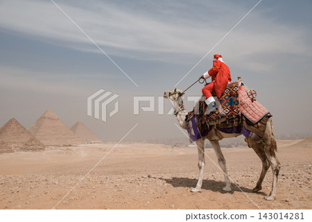 Wide shot back view of costumed Santa Claus on decorated camel facing all three Giza pyramids across sandy Egyptian desert plateau ideal for holiday Wide shot back view of costumed Santa Claus on decorated camel facing all three Giza pyramids across sandy Egyptian desert plateau ideal for holiday 143014281