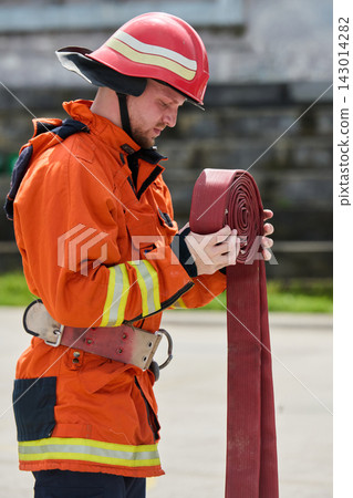 Firefighter In Orange Protective Uniform Rolling A Fire Hose While Wearing Helmet And Safety Gear Firefighter In Orange Protective Uniform Rolling A Fire Hose While Wearing Helmet And Safety Gear 143014282