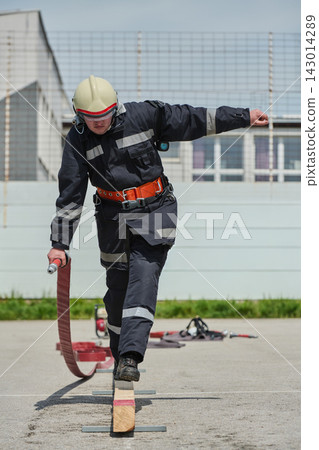 Firefighter Training Balancing On Beam While Carrying Hose During Outdoor Safety Drill Exercise Firefighter Training Balancing On Beam While Carrying Hose During Outdoor Safety Drill Exercise 143014289